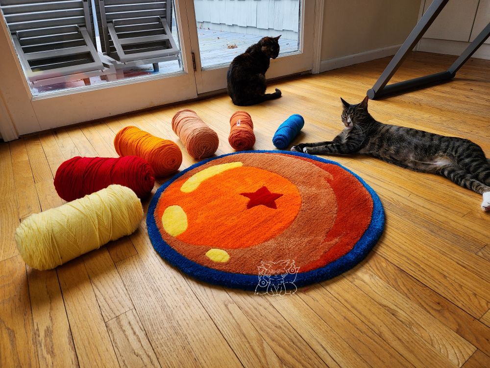 A dragon ball on a wooden floor in the form of a tufted textile artwork. With the tufted work, there are 2 cats, one torti and one tabby, and the corresponding yarn colors (yellow, red, few shades of orange, and blue).