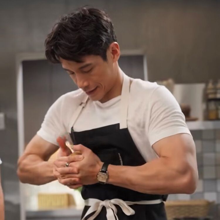 Manny Jacinto wearing a white tee with a black apron. He is in a kitchen looking down at a food object that he is trying to forcefully crush between his hands. So of course, his arms are flexed.  How dare. 