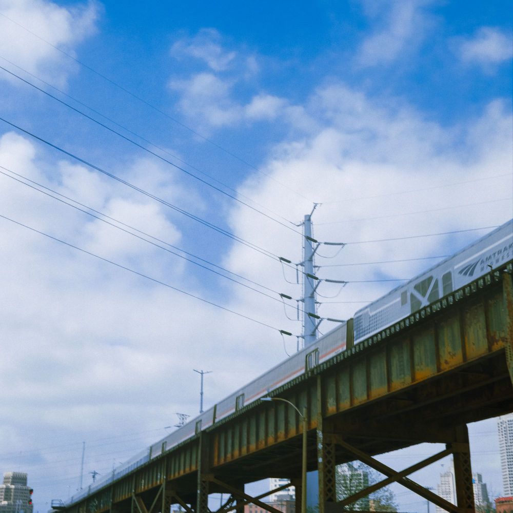 An Amtrak train going across a green and rusting bridge with a vibrant mostly cloudy sky in the background. Shot on Kodak Gold with a Minolta Autocord.