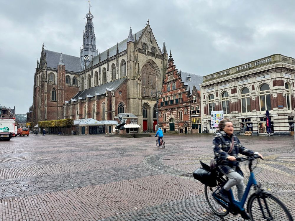 A photograph of the central Square and a large cathedral people are assuming past on bikes, especially the lady in the foreground she’s going places.
