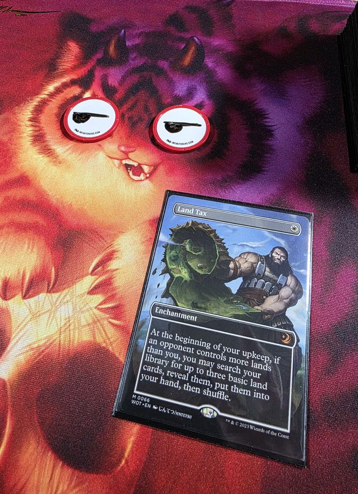 A Magic Land Tax on a playmat that has a cat on it. On the eyes of the cat are two InfiniTokens dry erase counters that have eyeballs drawn on them.