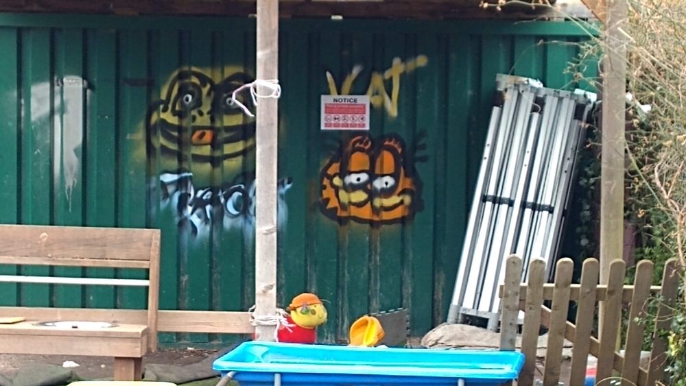 Photo of a green, corrugated steel wall in the garden area of a daycare nursery. On the wall are blurry, menacing, spray-painted faces of cartoon cat Garfield and a frog (or possibly a toad). Some worn children's toys and rough wooden furniture are visible in the foreground.