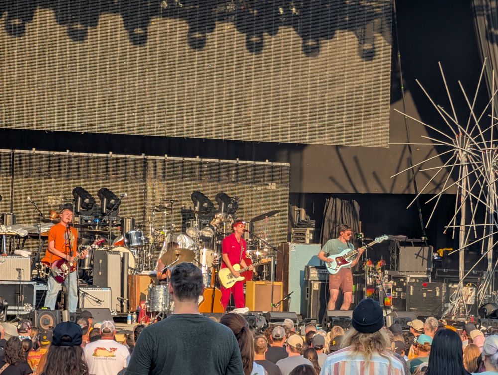 The band Friko playing a show at Stage AE in the low Pittsburgh evening sun. Two guitar players (with one singing lead vocals), a bass player, and a drummer, jamming.