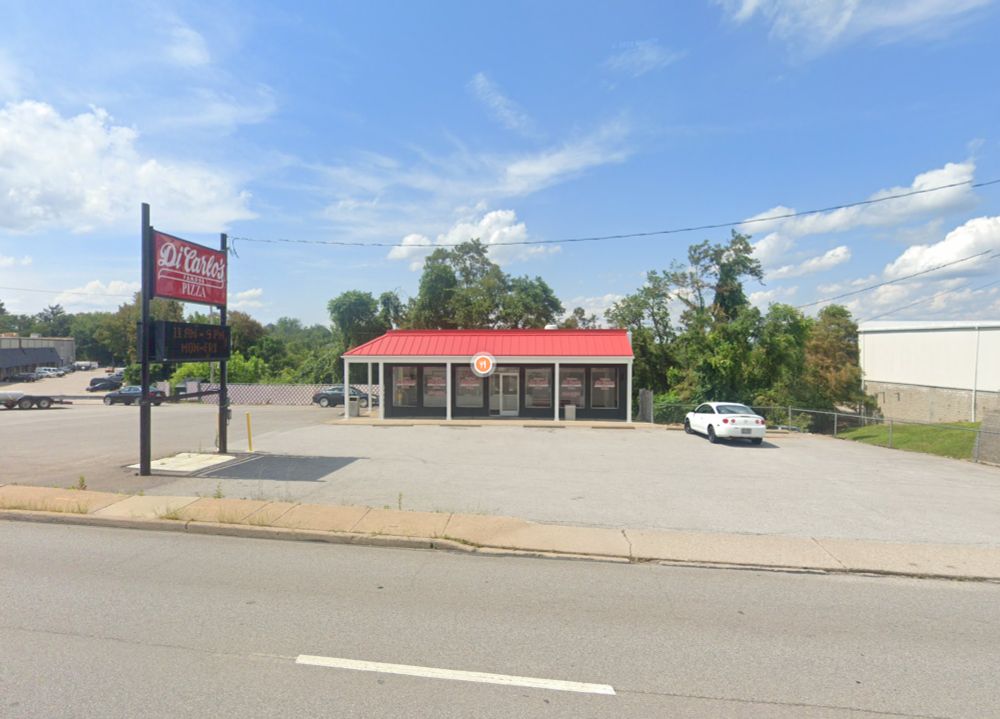 The modern/current view of the DiCarlo's pizza at 4531 Sunset Blvd, Steubenville, OH. The picture shows the same view as depicted in the first picture, with the building altered to have a red metal roof, no arches, and an internally-lit, printed red sign. The parking lot and trees (and sky, incidentally), remain the same as the other picture.