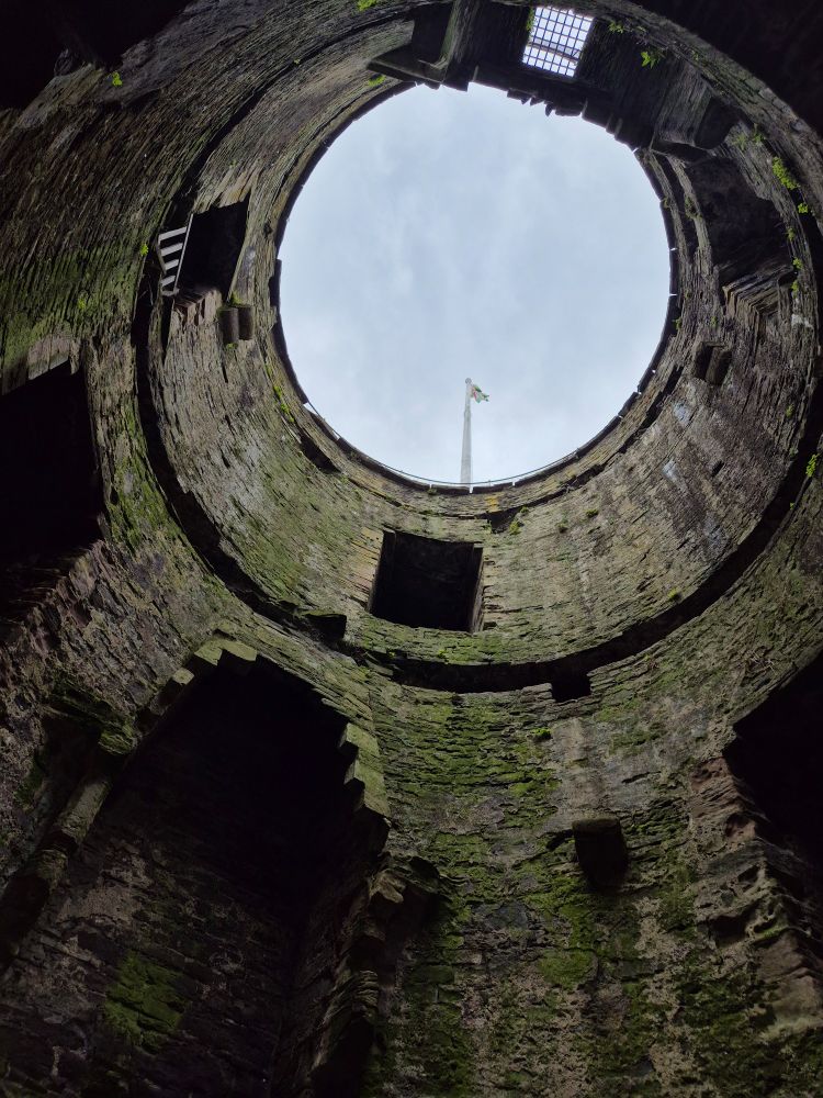 Inside a ruined castle tower, looking up through the missing levels to a flag pole and the cloudy sky.