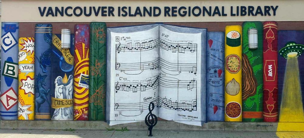 This image shows a colorful mural on the exterior wall of the Vancouver Island Regional Library. The mural features a series of painted books with unique designs. The central book is open and displays musical notes and annotations, celebrating the diverse cultural and educational resources available at the library.