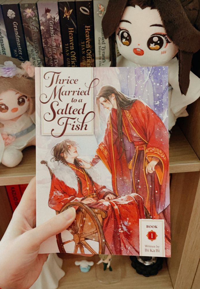 a picture of a hand holding a copy of "thrice married to a salted fish" (vol.1) by bi ka bi. on the background there's a bookshelf with a few danmeis and two xie lian plush dolls.