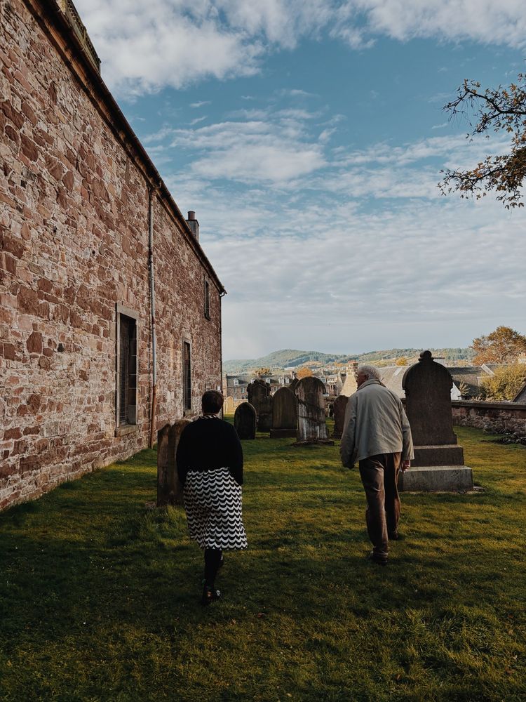 Two figures walk on dark green grass of a graveyard, towards the centre of the image - their backs facing the photographer. To the left of the photo is the wall of the church which is brown and yellow sandstone blocks. The woman walks slightly behind the man, wearing a white and black striped dress and black coat and has short hair. The man wears brown trousers and beige coat.  The sky is blue with thin whispy clouds