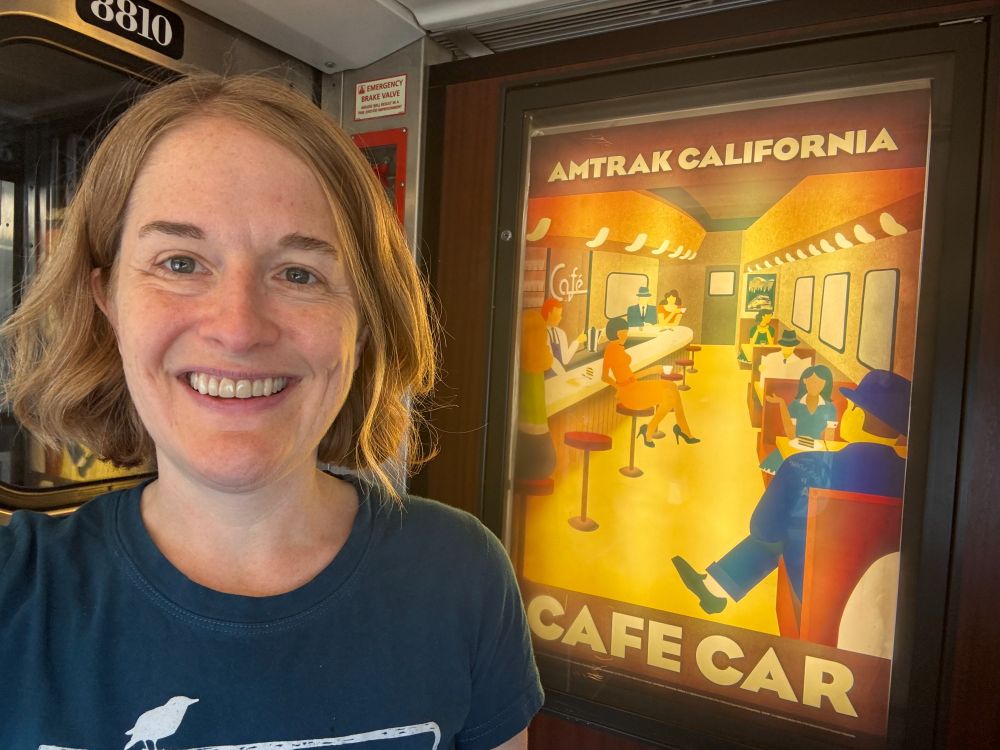 Me smiling on Capitol Corridor, with Amtrak California Cafe Car poster behind me.