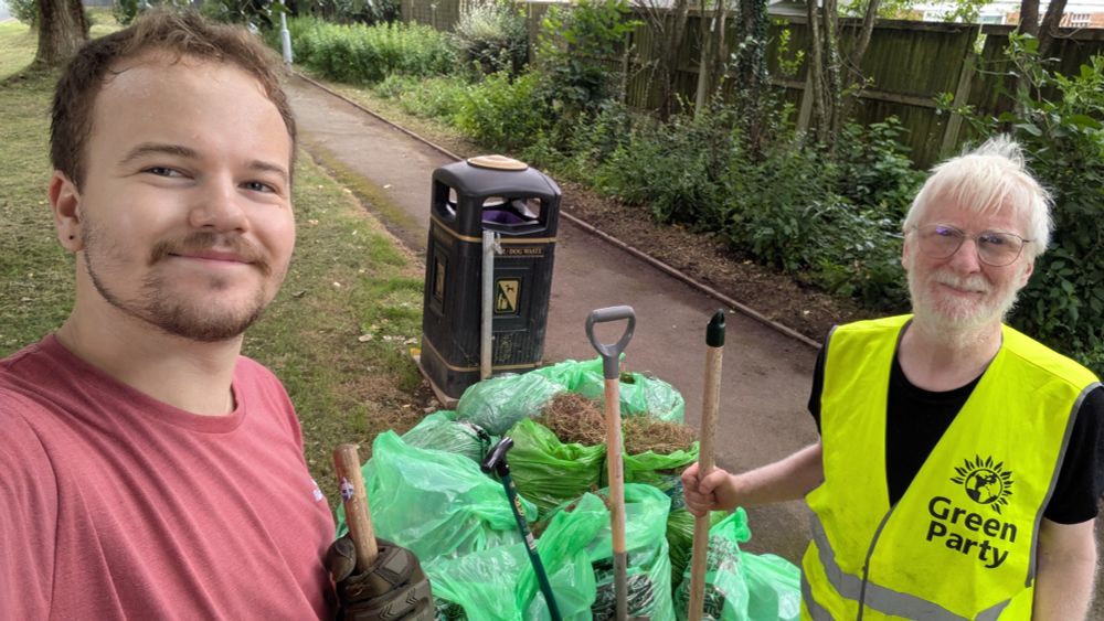 A young man with brown hair and beard (Hamish) and an older man with white hair and beard  and wearing a high-vis vest with the Green Party logo (Andrew), stand smiling with gardening tools, in front of 15 bags of weeds cleared from the pathway behind. 