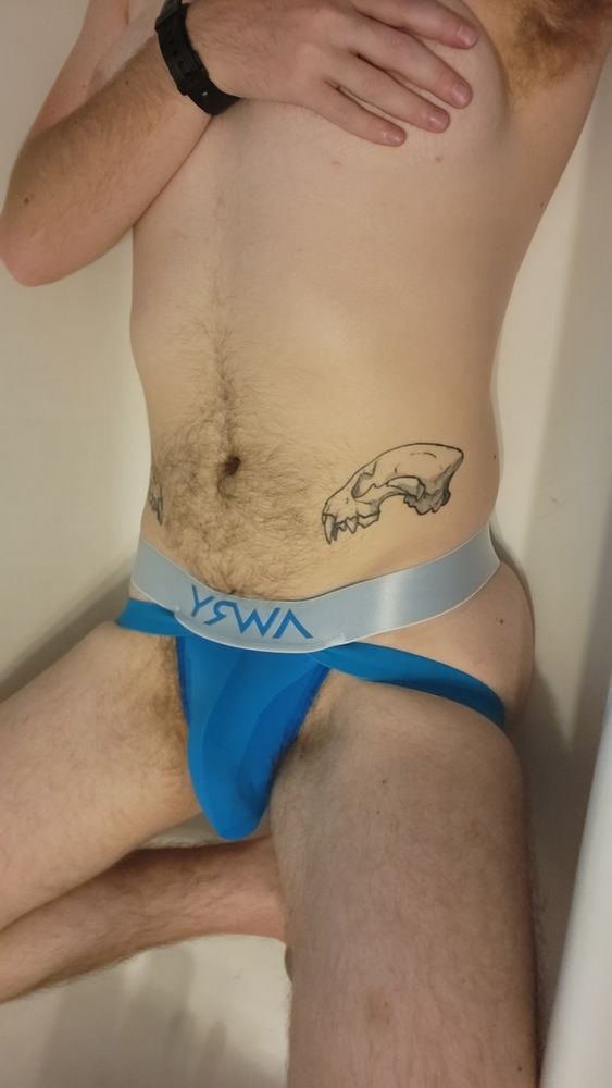 A shot of Blue from his knee to his shoulders. I'm laying in the bathtub in nothing but a bright-blue and silver jockstrap. My hip tattoo is visible (a hyena skull), as well as my tummy hair. I'm covering my chest with my hand.