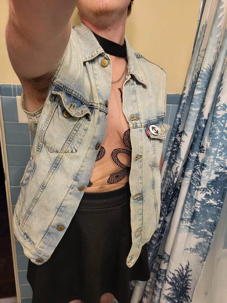 Blue from his knees to his chin. He's wearing a black short skirt, his black Thistle And Spire Medusa bodysuit, and a light-wash denim vest with a switch ↕️ pin on the pocket.