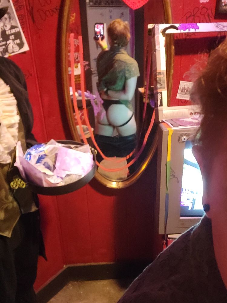 A slightly blurry mirror photo of Blue in a venue bathroom. His black pants are pulled down to his his mid-thigh and one arm is behind his back, holding his green lace shirt out of the way and showing a 3/4 shot of his new jock-thong.