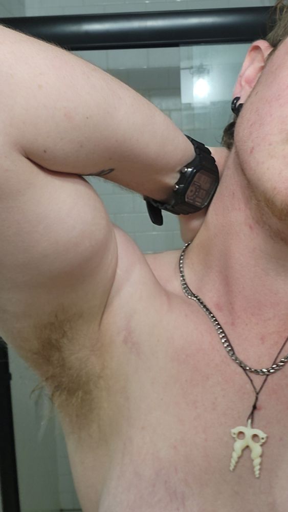A shot showing Blue from his sternum to his ear, showing just the left side of his body. He has his arm up and his hand tucked behind his neck to show off his bicep. He is wearing a watch, a stainless steel miami cuban chain, and a pendant depicting a symbol of Nyami-Nyami, the Zambezi river god.