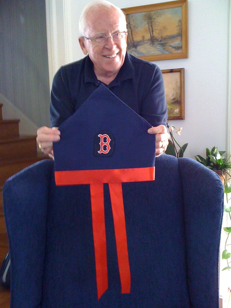An older caucasian man in a blue shirt holds a navy blue and red bishop's miter with a Red Sox B logo on it, resting atop a blue padded chair. Behind him are stairs and a couple of framed paintings of landscapes, as well as some house plants