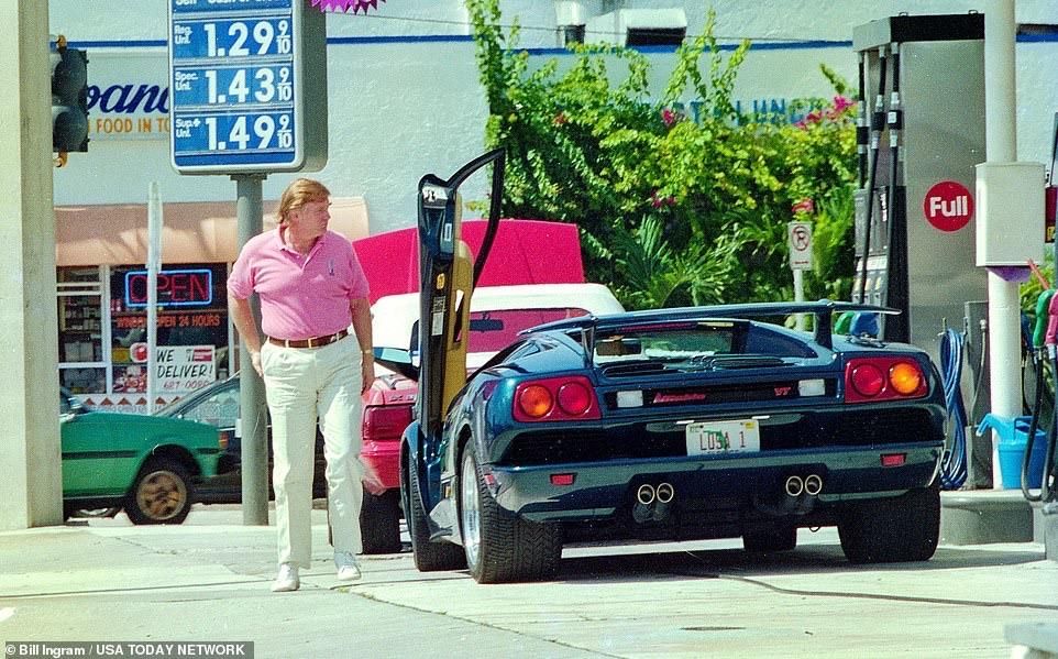 A man in a pink polo shirt and white pants stands next to a black Lamborghini at a gas station. The car's door is open, and gas prices are visible on a sign in the background. A green vehicle and a store can be seen in the background. 