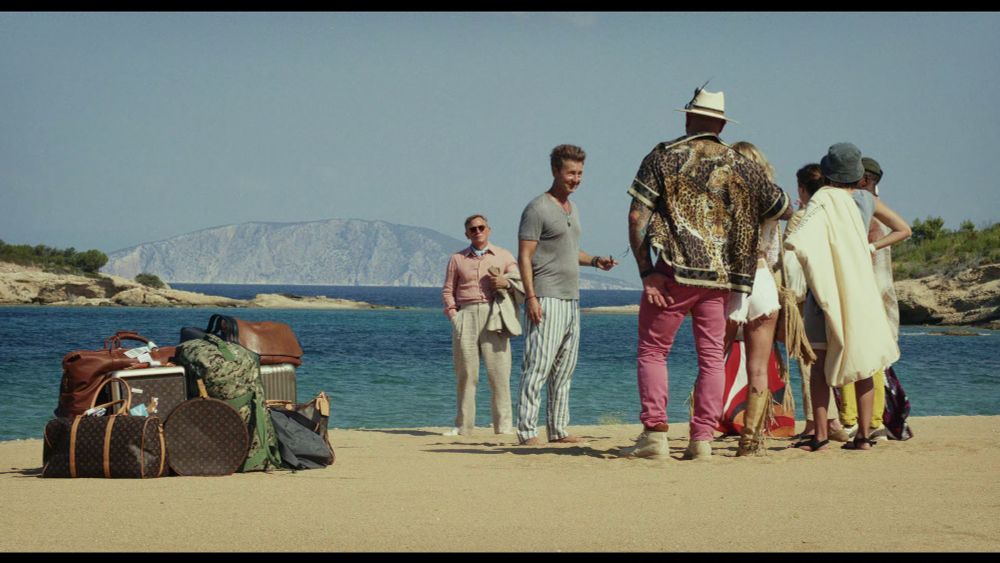 A joyfully composed shot of the cast on the beach, the cast and their luggage on either side. Daniel Craig's Benoit Blanc occupies the center, in the background.