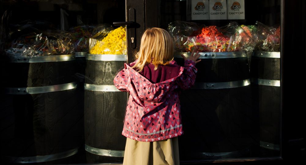 Child up against glass doors. Piles of candy on a bunch of barrels on the other side of the doors.