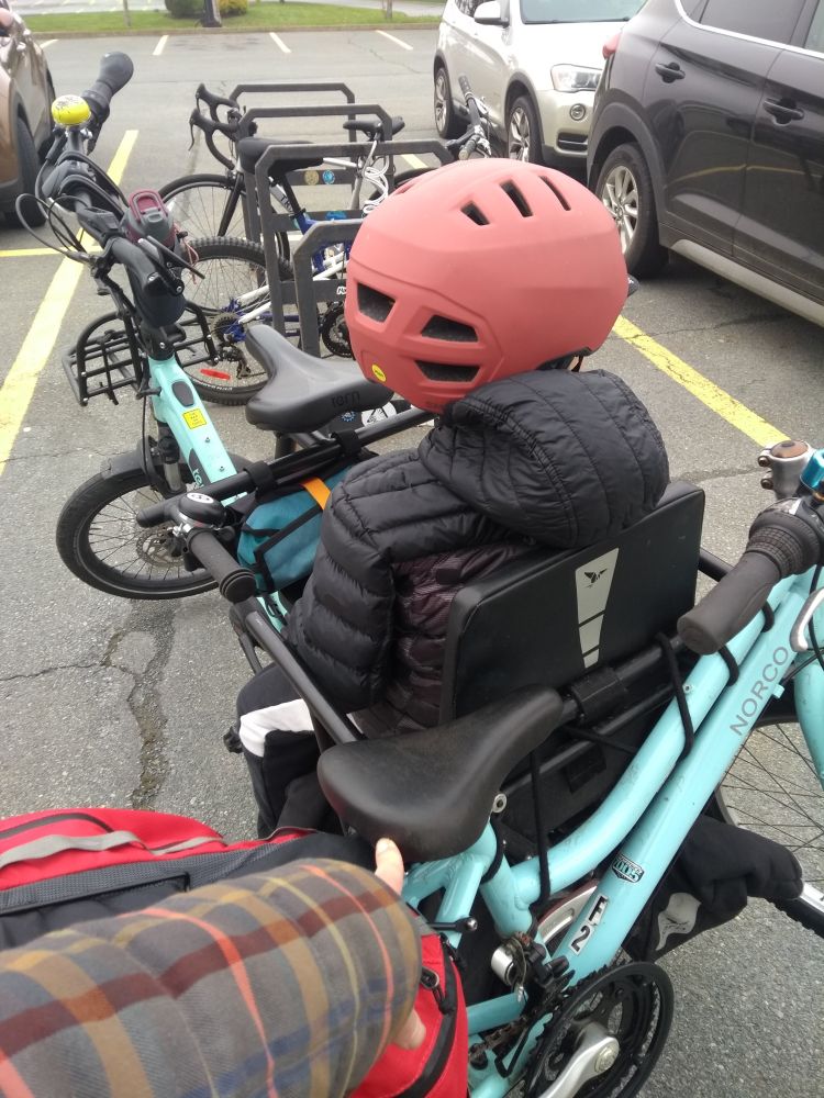 Child sitting on the back of a long tailed e-cargo bike. His 20" wheel bike strapped to the back. In the background is a bike rack in a large parking lot with cars parked beside it.
