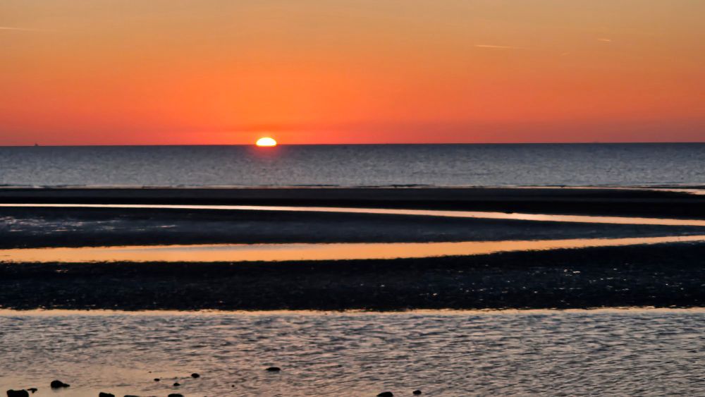 Sun setting over the sea.  Orange red sky, blue water, and streaks of sand and water in foreground because tide has gone out. 