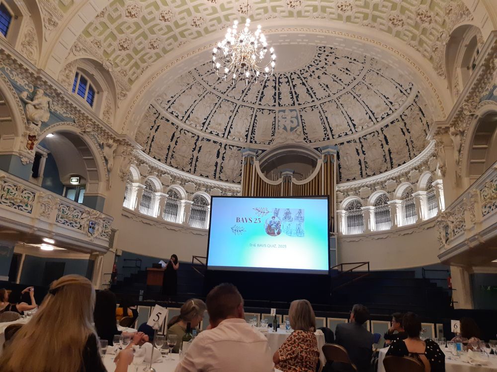 Ornate plaster domed roof with chandelier and people in foreground seated at round dinner tables plus big screen saying BAVS 2025