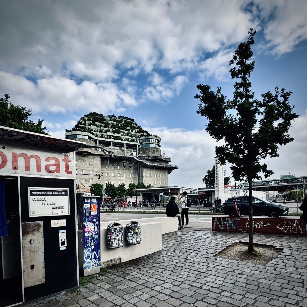 Urbanes Stadtbild mit einem großen Betongebäude im Hintergrund, das mit Pflanzen auf Dach und Terrassen begrünt wurde. Die Struktur wirkt wie ein umgenutzter Bunker oder Industriebau, nun modernisiert für Wohn- oder Geschäftszwecke. Im Vordergrund befindet sich ein öffentlicher Bereich mit einem Baum, gepflastertem Weg und einem kleinen Automatenhäuschen. Zwei bunt dekorierte Mülleimer stehen daneben, einige Menschen sind zu sehen. Der Himmel ist teils bewölkt. Die Szene zeigt den Kontrast zwischen historischer Architektur und zeitgenössischer Stadtentwicklung.