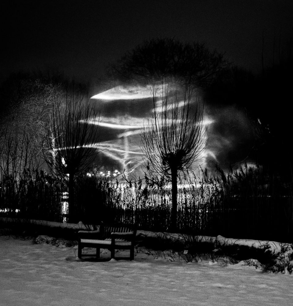 Ein nächtliches Schwarz-Weiß-Foto zeigt eine verschneite Landschaft mit einer leeren Bank im Vordergrund, kahlen Bäumen und hohem Gras. Im Hintergrund durchschneiden helle Lichtstrahlen den Nebel und erzeugen eine dramatische, fast surreale Atmosphäre. Die Szene wirkt ruhig und geheimnisvoll, als Teil einer künstlerischen Lichtinstallation.