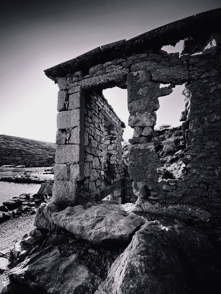 Schwarz-weiß Foto einer aus Naturstein erbauten Ruine auf der Insel Krk. Im Hintergrund scheint die Sonne auf das alte Bauwerk, seitlich sind eine kleine Bucht der Adria und eine Anhöhe zu erkennen.