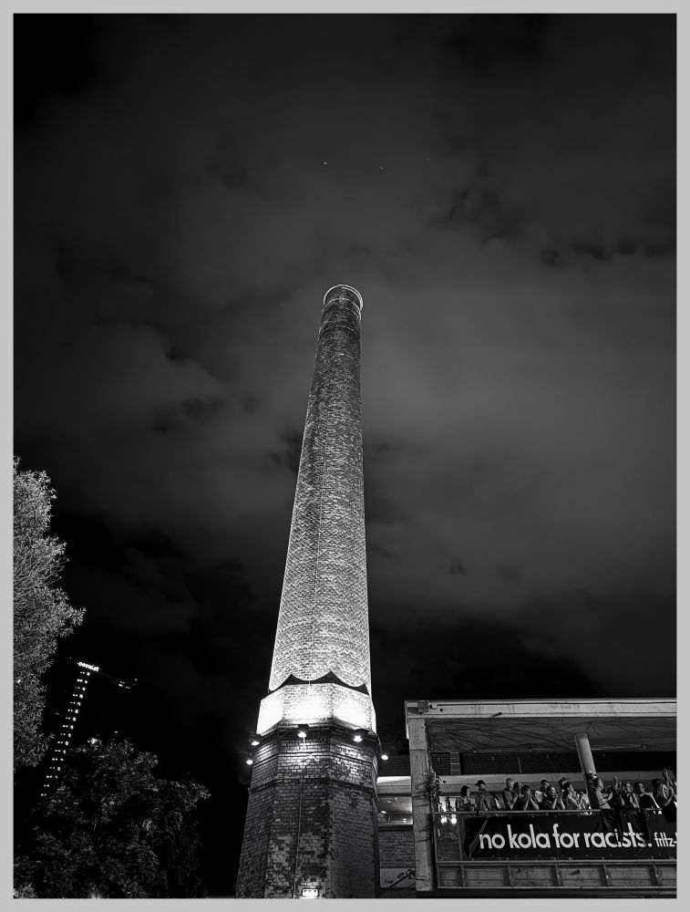 Das Bild zeigt einen hohen, beleuchteten Schornstein bei Nacht, aufgenommen in Schwarz-Weiß. Der Schornstein ist aus Ziegeln gebaut und steht neben einem Gebäude, auf dessen Balkon sich eine Gruppe von Menschen befindet. An der Balkonbrüstung hängt ein Banner mit der Aufschrift "no kola for racists #fitz". Im Hintergrund sind Bäume und ein bewölkter Himmel zu sehen, während links ein Teil eines weiteren Gebäudes sichtbar ist. Die Szene vermittelt eine urbane Atmosphäre mit einem protestartigen Element durch das Banner.