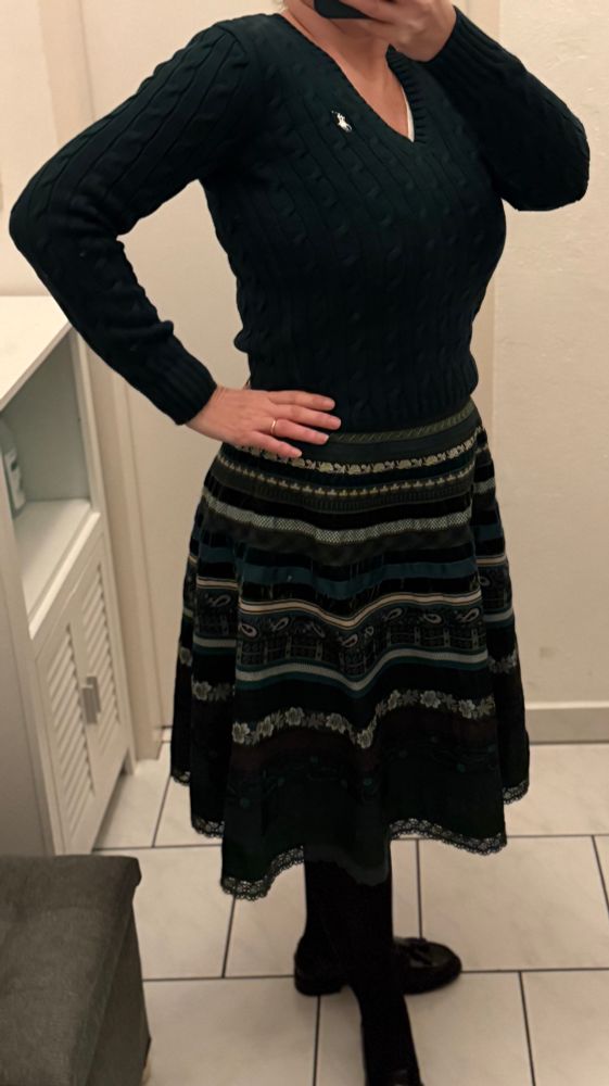 Ein aus grünen, schwarzen und goldenen Bänder bestehender Rock. Dazu ein dunkelgrüner Pullover mit V-Ausschnitt im Zopfmuster