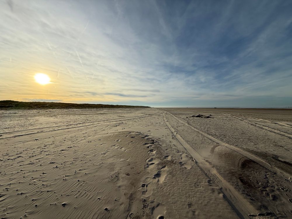 Foto des Strandes auf Norderney. Links die tief stehende Sonne über weit entfernten Dünen. Am Himmel sind ein paar Wolken. 