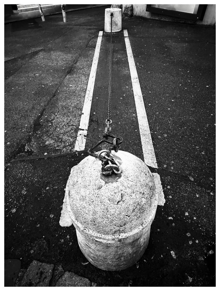 Das Schwarz-Weiß-Foto zeigt einen Betonpoller auf einem asphaltierten Untergrund, der mit einer dicken Kette und einem Vorhängeschloss an einem zweiten, kleineren Betonpfosten in der Ferne gesichert ist. Die Kette verläuft entlang einer weißen Markierungslinie auf dem Boden. Der Poller im Vordergrund ist rund, verwittert und weist Risse sowie Abplatzungen auf. Der Asphalt ist ebenfalls abgenutzt, mit Rissen und kleinen Schmutzpartikeln. Im Hintergrund sind unscharf weitere Betonstrukturen und ein Gebäude zu erkennen.