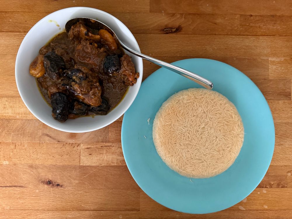 Das Bild zeigt eine Mahlzeit, die auf einem Holztisch serviert wird. Es gibt eine weiße Schüssel mit einem dunklen, würzigen Eintopf, der Fleisch und Pflaumen und Marillen enthält, zusammen mit einer Kelle. Daneben steht ein blauer Teller mit einer Portion crispy Reis