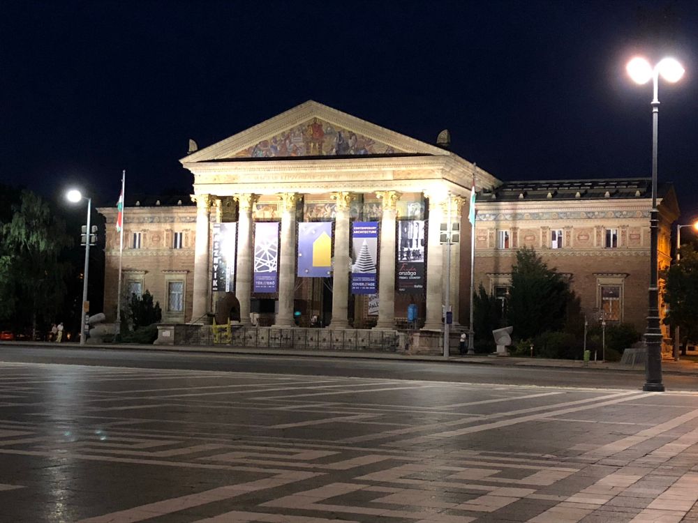 Auf dem Bild sieht man ein imposantes klassizistisches Gebäude bei Nacht, das von mehreren Laternen und Scheinwerfern angestrahlt wird. Es handelt sich um das Kunsthalle (Műcsarnok) in Budapest, Ungarn, ein Ausstellungsgebäude für zeitgenössische Kunst. Das Bauwerk zeichnet sich durch seine eindrucksvolle Tempelfassade mit sechs Säulen aus, die einen Dreiecksgiebel tragen. Der Giebel ist mit farbigen Mosaiken geschmückt. Zwischen den Säulen hängen große Banner mit Informationen zu aktuellen Ausstellungen. Im Vordergrund ist ein weitläufiger Platz mit geometrisch gemustertem Steinboden zu sehen. Die nächtliche Beleuchtung hebt die architektonischen Details des Gebäudes besonders stark hervor.