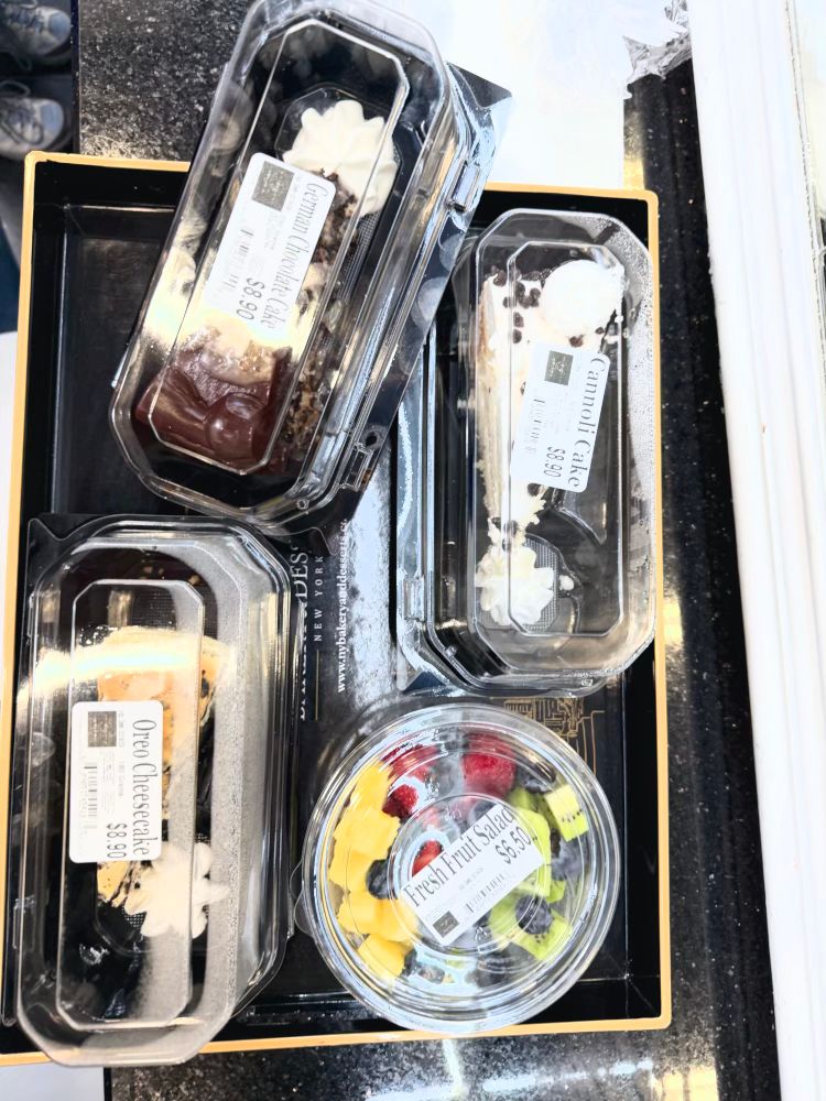 A selection of packaged desserts and fruit salad displayed on a black tray. The visible items include:
	1.	A “German Chocolate Cake” slice in a plastic container, priced at $8.90.
	2.	A “Cannoli Cake” slice in a plastic container, priced at $8.90.
	3.	An “Oreo Cheesecake” slice in a plastic container, priced at $8.90.
	4.	A “Fresh Fruit Salad” in a clear plastic cup, priced at $6.50, containing a mix of pineapple, kiwi, blueberries, raspberries, and strawberries.

The desserts and fruit are neatly packaged, placed on a glossy black counter.