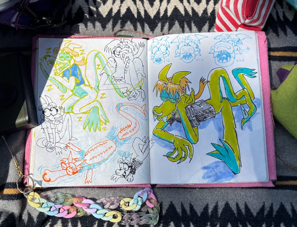 a sketchbook spread photographed on a blanket surrounded by pen bags and a plastic chain. It’s a spread of iggys, Iggy sleeping, iggy sitting cutely, a submerged gator iggy, three iggys drawn with a progressively wider angle, and a colored marker drawing of iggy sitting in an impossible pose i couldnt replicate myself