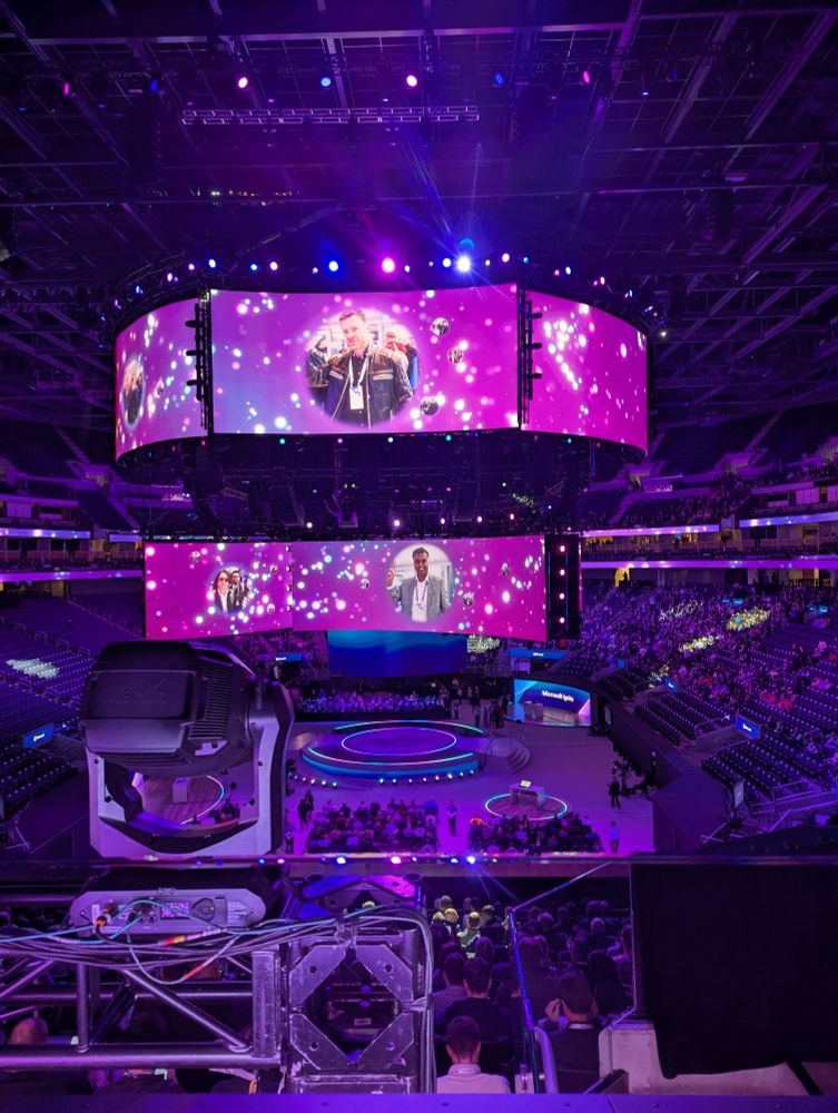 Alt text: A large-scale tech conference, Microsoft Ignite, held in an indoor arena with tiered seating filled with attendees. The central circular stage is illuminated in vibrant purple and blue hues, surrounded by professional lighting rigs. Above the stage, a massive circular screen displays animated sparkles and portraits of two individuals, suggesting keynote speakers or honorees. A control panel is visible in the foreground, emphasizing the event’s high production value and immersive audiovisual setup.