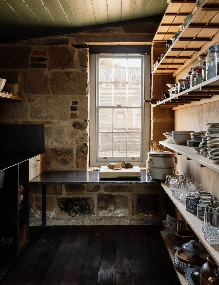 A view of a renovated kitchen in an old stone cottage. Wooden shelves line one wall; a modern countertop runs beneath a window.