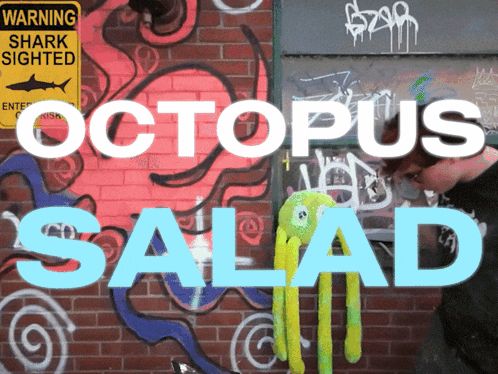 a man stands in front of a brick wall with graffiti and a sign that says warning shark sighted octopus salad