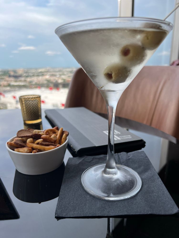 A martini with three olives and some bar snacks on a table with large windows and a city view in the distance. 