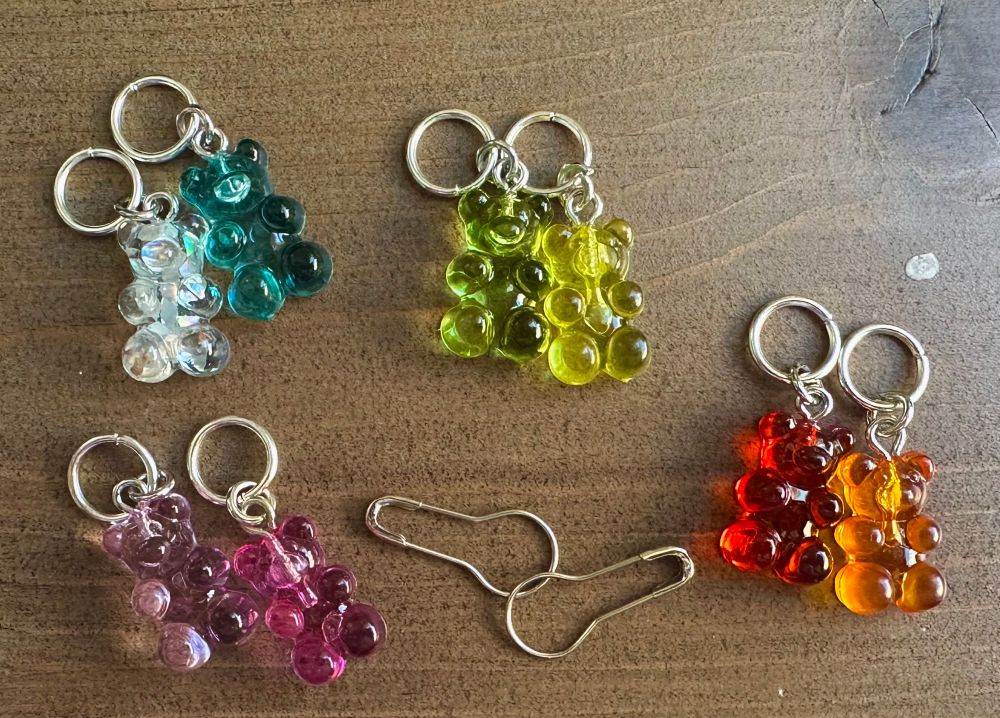 Semi transparent, gummy bear stitch markers in various colors with 10mm rings laying on a wooden backdrop with two silver bulb pins.  The gummy bears are arranged to show the different color sets… Sparkly white and blue, green and yellow, red and orange, & purple and pink.