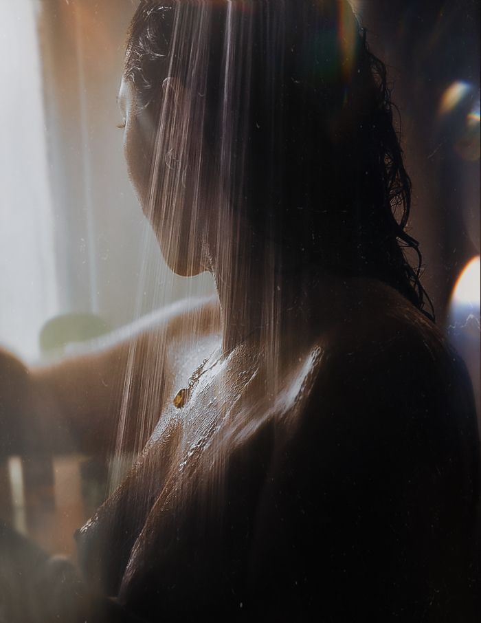 A fine art nude photograph of a woman standing under a warm shower, captured from behind and in profile. The image is softly lit with natural sunlight streaming in from the left side of the frame, casting a golden, ethereal glow throughout the scene. Water flows down her body in delicate streaks, catching the light and emphasizing the texture of her wet skin. Her dark hair is slicked back, and her face is turned gently to the side, mostly in shadow, creating a sense of privacy and introspection. A single pendant necklace rests on her chest, glinting faintly in the light. The background is softly blurred, contributing to the moody, intimate atmosphere of the photograph. The overall tone is warm, soft, and serene, blending sensuality with calm solitude.