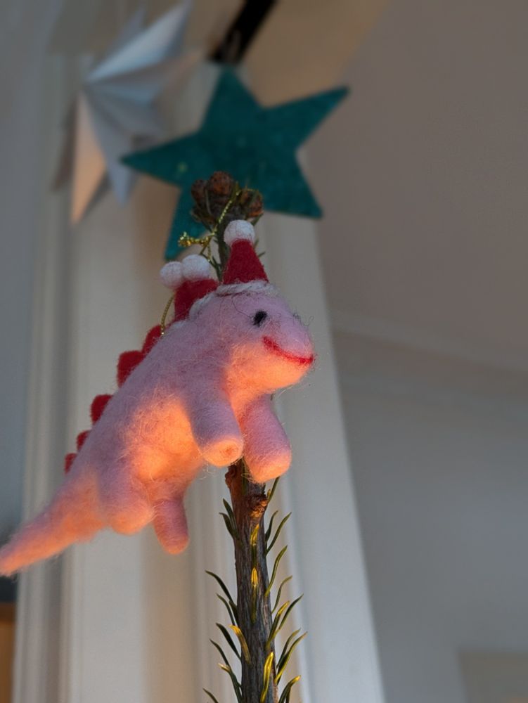 Ein lächelnder, pinker Dinosaurier mit drei Weihnachtsmannmützen an der Tannenbaumspitze.