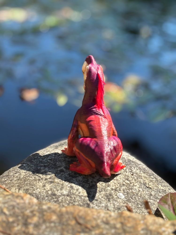 a back veiw of the model photographed on a rock in front of some lillies. It is a Pettable Gorgon model, which is a slightly chibi gorgonopsid going blep and squeenching up its eyes