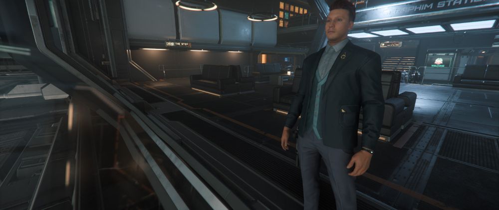 The image depicts a technologically advanced and futuristic indoor space, possibly part of a space station or high-tech facility. A character stands on the right, dressed in a teal suit with a matching tie and light blue shirt. The background features sleek metal and glass architecture, illuminated by modern, circular lights on the ceiling. To the left, there is an elevated walkway, with cautionary signage above lounge areas containing dark seating arrangements. The environment is spacious, with visible seating areas and different levels, creating a sense of depth.