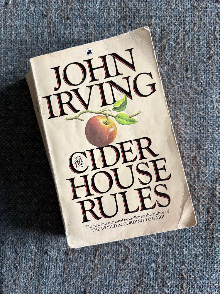 A paperback copy of Cider House Rules by John Irving laying on a grey couch cushion.