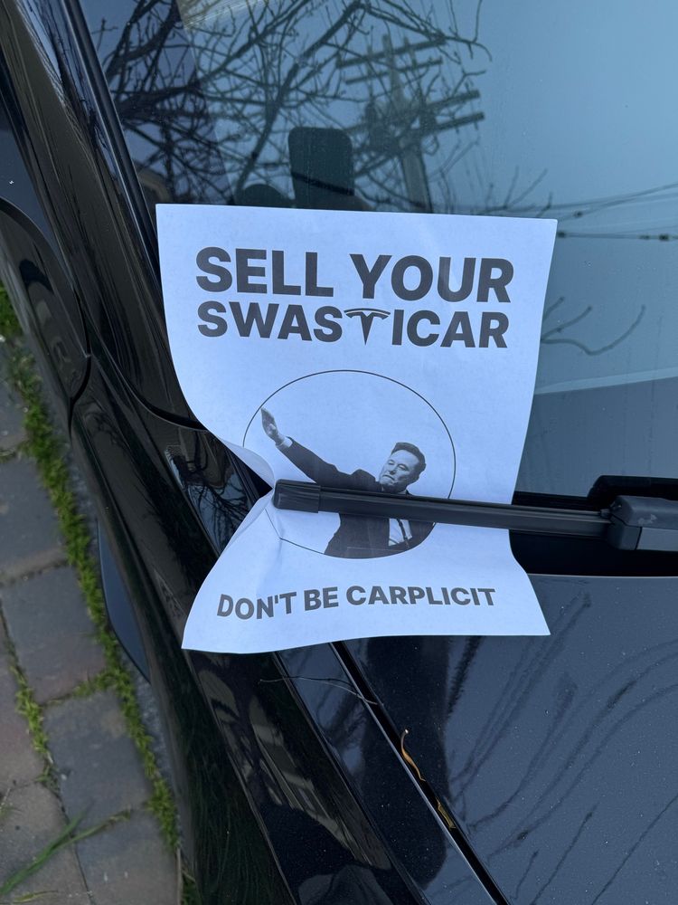 A flyer placed on the windshield of a black Tesla. The flyer reads:

SELL YOUR SWASTICAR
DON’T BE CARPLICIT

The “t” in “Swasticar” is the Tesla logo. In the middle of the flyer is the picture of Elon Musk  performing a Nazi salute. 