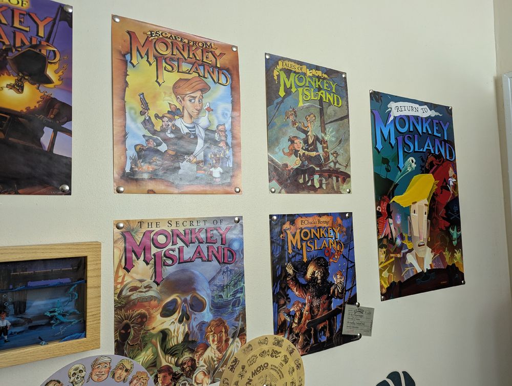 Two rows of Monkey Island game posters hanging on a wall. To the right is a somewhat larger Return to Monkey Island poster