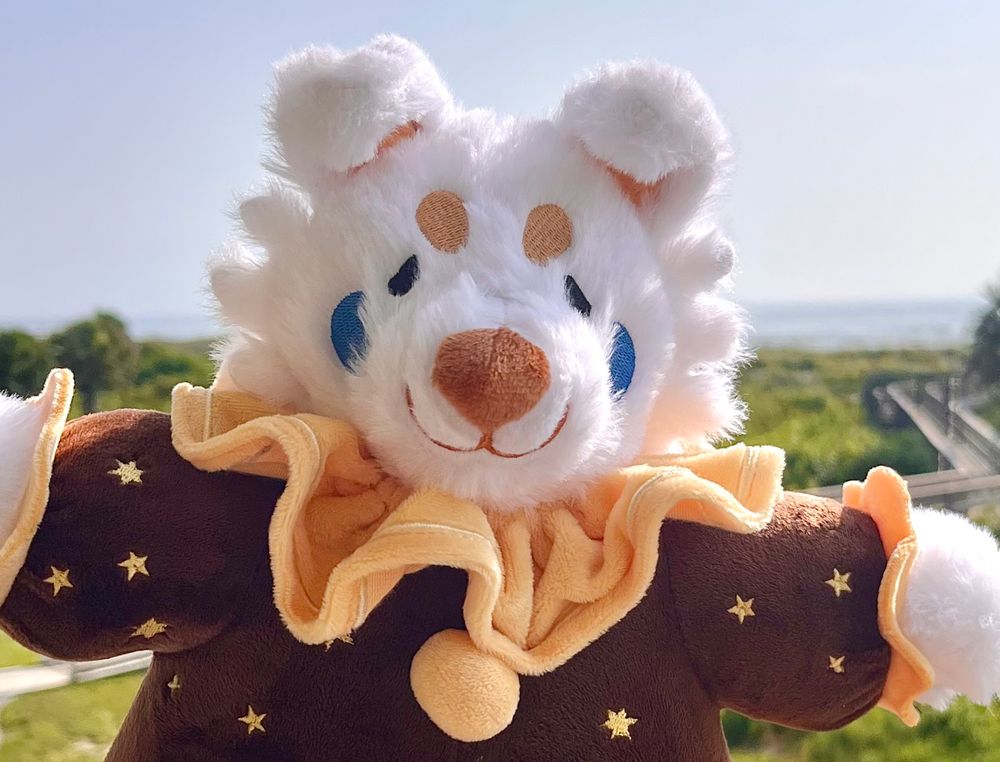Plush of a White dog with floppy ears dressed as a clown in a brown costume with yellow Pom Poms and collar with a star pattern