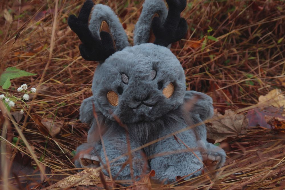 Grey rabbit plush with antlers and sharp teeth with orange blush and orange paws resembling a wolpertinger in a forest setting